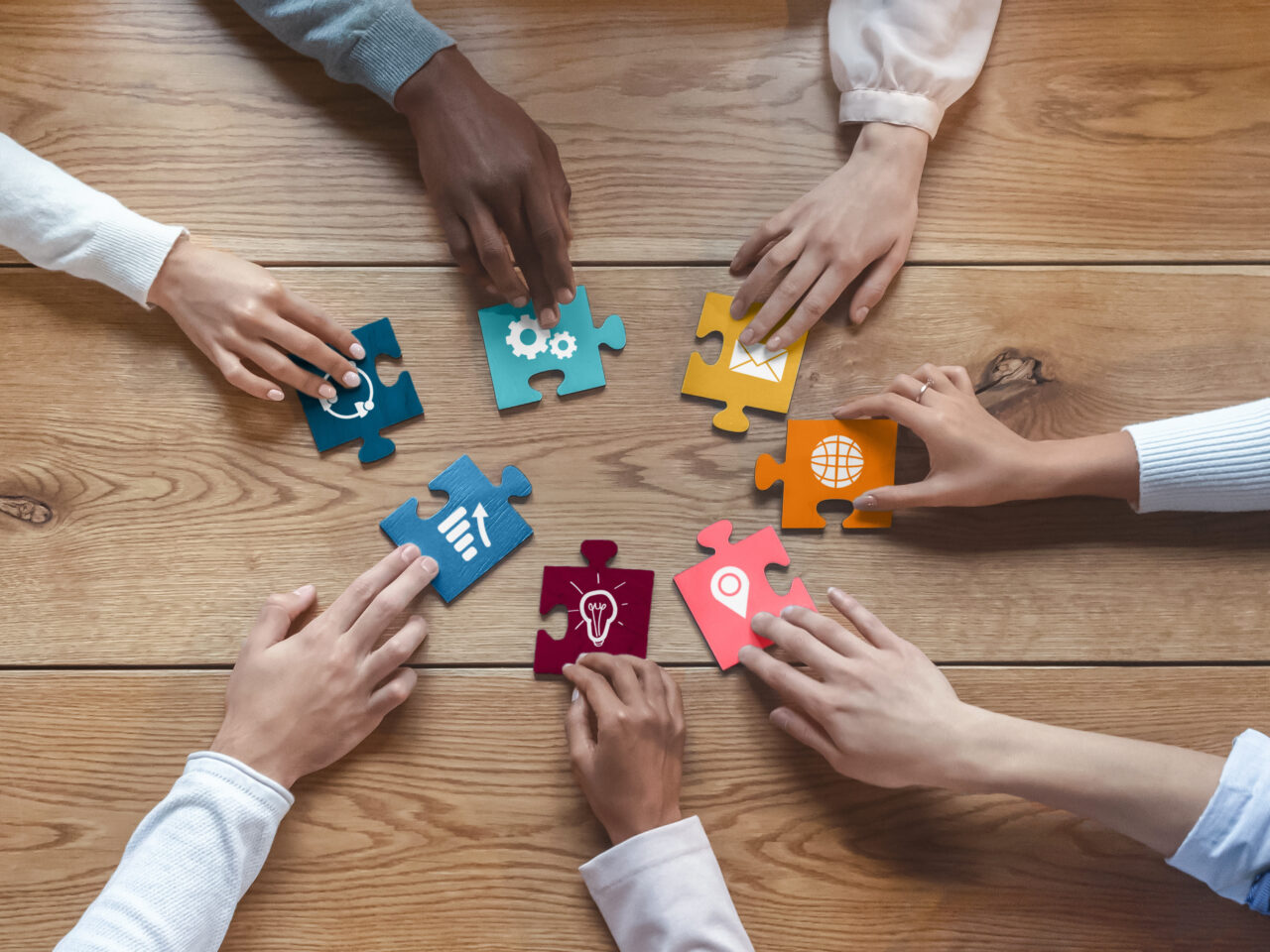 Hands of international coworkers sitting around table, putting colorful puzzles together, teamwork concept, top view